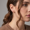 A close-up of a woman's ear wearing a delicate gold infinity-shaped dangle earring with two long hanging chains, against a soft gray background.