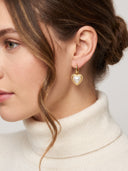 A close-up of a woman wearing a gold hoop earring with a heart-shaped white pearl pendant, paired with a cream turtleneck sweater.