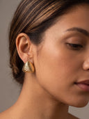 A close-up of a woman's ear and side profile wearing a textured, ribbed gold hoop earring against a neutral gray background.