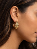 A close-up side profile of a woman wearing a gold crescent-shaped hoop earring with a twisted, layered design.