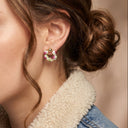 A close-up of a woman wearing a small gold hoop earring adorned with pink, green, and yellow crystals, paired with a denim jacket featuring a cream sherpa collar.