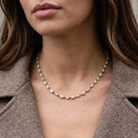 A close-up of a woman wearing a delicate gold tennis necklace with clear baguette-cut gemstones, styled with a brown blazer.