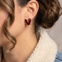 A close-up of a woman wearing a small heart-shaped stud earring with a deep red enamel center and gold-tone border.