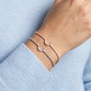 Two delicate crescent moon bracelets, one in yellow gold and one in silver, both set with small clear stones, worn on a person's wrist over a light blue knit sweater.