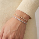 A close-up of a person's wrist wearing three delicate bracelet styles: a gold chain with a star-shaped charm, a silver chain with a star-shaped charm, and a clear stone tennis bracelet. The person is wearing a beige knit sweater.