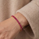 A close-up of a gold bracelet adorned with small, vibrant pinkish-red gemstones worn on a person's wrist, set against a soft beige knit sweater.