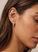 A close-up of a woman's ear wearing a small gold huggie hoop earring adorned with tiny white pearls, set against a soft beige background.