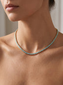A close-up of a model wearing a delicate gold tennis necklace adorned with small round turquoise-blue stones.