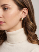 A close-up of a woman wearing a small square gold stud earring with a textured, ribbed surface, paired with a cream-colored ribbed turtleneck sweater.