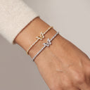 Two delicate butterfly bracelets, one in yellow gold and one in silver, both set with small clear stones, shown worn on a person's wrist against a soft beige background.