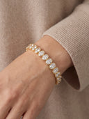 A gold tennis bracelet with round clear crystals, worn on a person's wrist against a beige ribbed knit sweater.