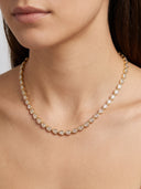 A close-up of a model wearing a delicate gold tennis necklace with clear teardrop-shaped stones.