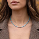 A close-up of a woman wearing a delicate silver tennis necklace with small, round turquoise-blue stones, styled with a brown blazer.