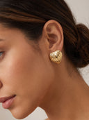 A close-up of a woman's ear wearing a textured, ribbed gold heart-shaped stud earring against a soft beige background.