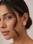 A close-up of a woman wearing a textured gold heart-shaped stud earring, shown against a soft beige background.