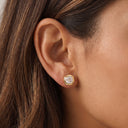 A close-up of a model's ear wearing a small gold stud earring with a cushion-cut clear gemstone.