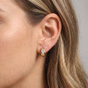 A close-up of a woman's ear wearing a small gold huggie hoop earring with a clear marquise-cut gemstone.