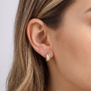 A close-up of a woman's ear wearing a small gold huggie hoop earring with a clear gemstone, shown against her hair and skin.