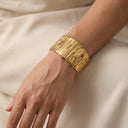 A wide, chunky gold cuff bracelet with a textured, hammered finish, shown worn on a person's wrist against a soft beige fabric background.