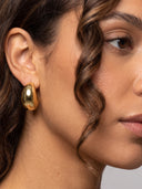A close-up of a woman's ear wearing a chunky, polished gold teardrop-shaped hoop earring.