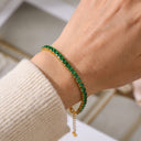 A close-up of a person's wrist wearing a gold tennis bracelet set with green rectangular gemstones, paired with a cream-colored knit sweater sleeve.