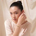 A woman with her hair in a bun wears a delicate gold chain bracelet on her wrist, posing against a soft beige draped fabric background.