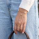 A close-up of a person's hand wearing a delicate gold chain hand bracelet with star and pearl details, paired with light blue jeans and a white ribbed sweater cuff.