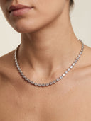 A close-up of a model wearing a delicate silver tennis necklace with clear teardrop-cut gemstones against a warm beige background.