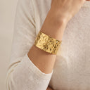 A wide, hammered gold cuff bracelet worn on a person's wrist, paired with a cream-colored knit sweater.