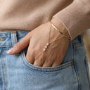 A close-up of a person's hand in a jeans pocket wearing a delicate gold chain hand bracelet with small clear stones, paired with a gold bangle and a beige ribbed sweater.