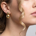 A close-up of a woman's ear wearing a gold drop earring with two polished spherical beads, shown against a light gray background.