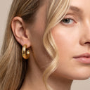 A close-up of a model's ear wearing a thick, textured gold hoop earring against a soft beige background.