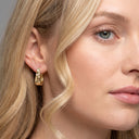 A close-up of a woman's ear wearing a small gold huggie hoop earring, with her blonde hair and face softly visible against a neutral gray background.