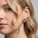 A close-up of a woman's ear wearing a gold-toned, knot-like stud earring against a soft gray background.