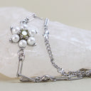 A silver-tone chain bracelet featuring a flower motif with white faux pearls and a central clear rhinestone, displayed on a white stone prop against a light beige background.