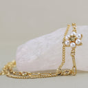 A gold chain bracelet with a flower-shaped pearl cluster and a central clear stone, displayed on a pale pink crystal against a soft beige background.