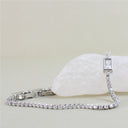 A silver tennis bracelet with clear rectangular and round stones, displayed on a white rock against a light beige background.