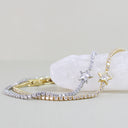Two delicate bracelets with star-shaped crystal pendants, one in yellow gold and one in silver, displayed on a white crystal cluster against a light beige background.