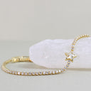 A gold tennis bracelet with clear cubic zirconia stones and a star-shaped crystal charm, displayed on a white crystal cluster against a light gray background.