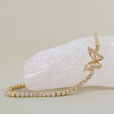 A gold tennis bracelet with clear pavé stones, featuring a bow-shaped pavé centerpiece, displayed on a white crystal cluster against a neutral beige background.