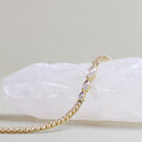 A gold tennis bracelet with marquise-cut clear stones, draped over a white crystal cluster on a neutral beige background.