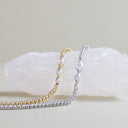 Two tennis-style chains draped over a white crystal display, one in yellow gold and one in silver, with clear gemstones set along the links against a soft beige background.