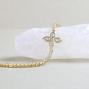 A gold chain bracelet with a four-petal flower charm set with clear stones, displayed on a white crystal block against a light beige background.