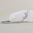 A silver tennis bracelet with a butterfly motif, set with clear pavé stones, displayed on a pale pink crystal against a neutral beige background.