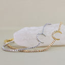 Two delicate crescent moon chain bracelets, one in yellow gold and one in silver, both paved with small clear stones, displayed on a white crystal cluster against a warm beige background.