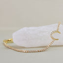 A gold tennis-style bracelet with a crescent moon charm, embellished with small clear stones, displayed on a white crystal against a light beige background.