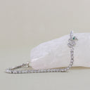 A silver tennis bracelet with a decorative spiral section featuring a small green heart-shaped gemstone, displayed on a white crystal block against a neutral beige background.