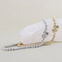 Two snake-shaped tennis bracelets with diamond-like cubic zirconia stones, one in yellow gold and one in white gold, each featuring a small green emerald accent, displayed on a white crystal block against a beige background.