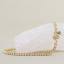 A gold tennis bracelet featuring a coiled snake motif adorned with clear stones and a small green gemstone at its head, displayed on a pale pink stone against a light beige background.