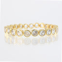 A gold eternity-style bracelet featuring round clear gemstones in bezel settings, shown on a clean white reflective surface.